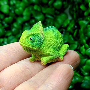 May include: A small, detailed figurine of a green chameleon with a textured skin. The chameleon has a distinctive head crest and is posed on a surface. The figurine is a vibrant shade of green, with hints of yellow and brown.