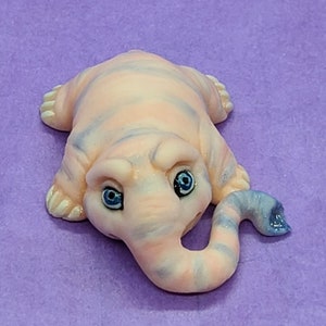 May include: A small, pink and blue striped figurine of a creature with a long, curled trunk and blue eyes. The figurine is made of a soft, pliable material and is lying on its side.