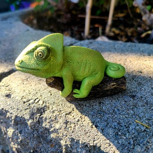 May include: A green chameleon figurine perched on a brown log. The chameleon has a textured surface and a large, round eye.