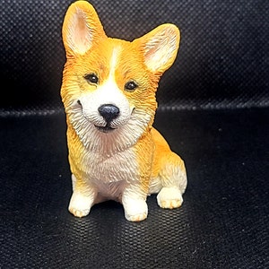May include: A small, ceramic figurine of a corgi dog. The dog is sitting with its front paws together and its back legs slightly spread. It has a white chest and white paws, with orange fur on its back and head. The dog has a friendly expression with black eyes and a black nose.