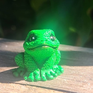 May include: A small green frog figurine with black eyes and a wide smile. The frog is sitting on a wooden surface.