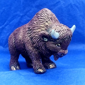 May include: A small, hand-painted ceramic figurine of a brown bison with a textured fur design. The bison has a silver horn and is standing on a blue surface.