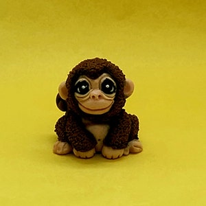 Pode incluir: Uma pequena figura de macaco marrom com olhos grandes e um rosto sorridente. A figura está sentada em uma superfície amarela.
