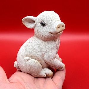 Op de afbeelding: Een kleine, gebroken witte varkensbeeldje met een glimlach. Het varkentje zit, met uitgestrekte voorpoten en opstaande oren. Het beeldje heeft een gedetailleerde vachttextuur en staat tegen een rode achtergrond.