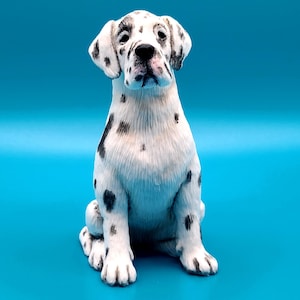 Può includere: Statuetta di cucciolo di dalmata bianco con macchie nere, seduto. Il cucciolo ha il naso nero ed è posto su uno sfondo blu brillante. Oggetto decorativo.