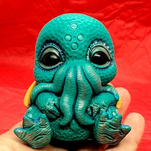 May include: A small, teal-colored, hand-painted ceramic sculpture of a sitting octopus with a textured surface. The octopus has large, black eyes and a yellow detail on its back.
