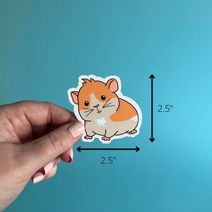 K&ouml;nnte beinhalten: Ein 6,35 cm x 6,35 cm gro&szlig;er Aufkleber mit einem Cartoon-Hamster mit orangefarbenem und wei&szlig;em Fell. Der Hamster schaut nach rechts und hat eine kleine, rosafarbene Nase und einen wei&szlig;en Mund.