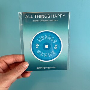 May include: A blue and white round sticker with the text "Muscle Mommy" and "25 KG" on it. The sticker is on a white card with the text "All Things Happy" and "stickers | magnets | stationery" on it.