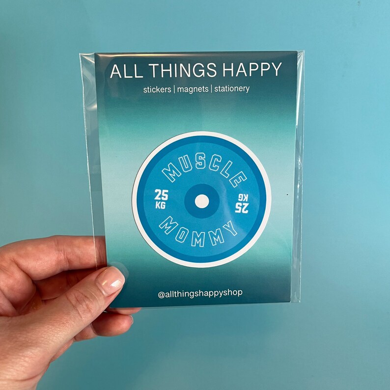 May include: A blue and white round sticker with the text "Muscle Mommy" and "25 KG" on it. The sticker is on a white card with the text "All Things Happy" and "stickers | magnets | stationery" on it.