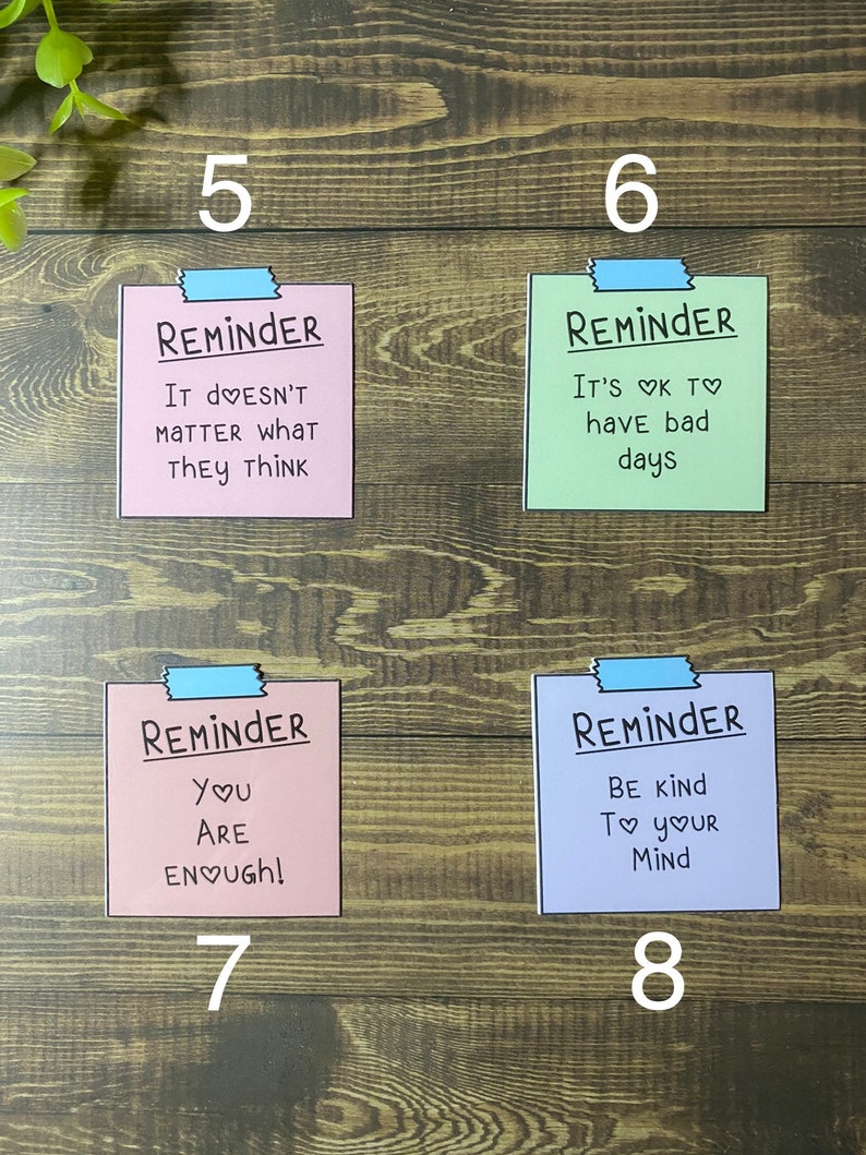 May include: Four pink, green, light blue and purple reminder notes with blue tape at the top. The notes have the word "Reminder" written in black at the top and inspirational messages in black with a pink heart symbol. The messages are: "It doesn't matter what they think", "It's ok to have bad days", "You are enough!" and "Be kind to your mind".