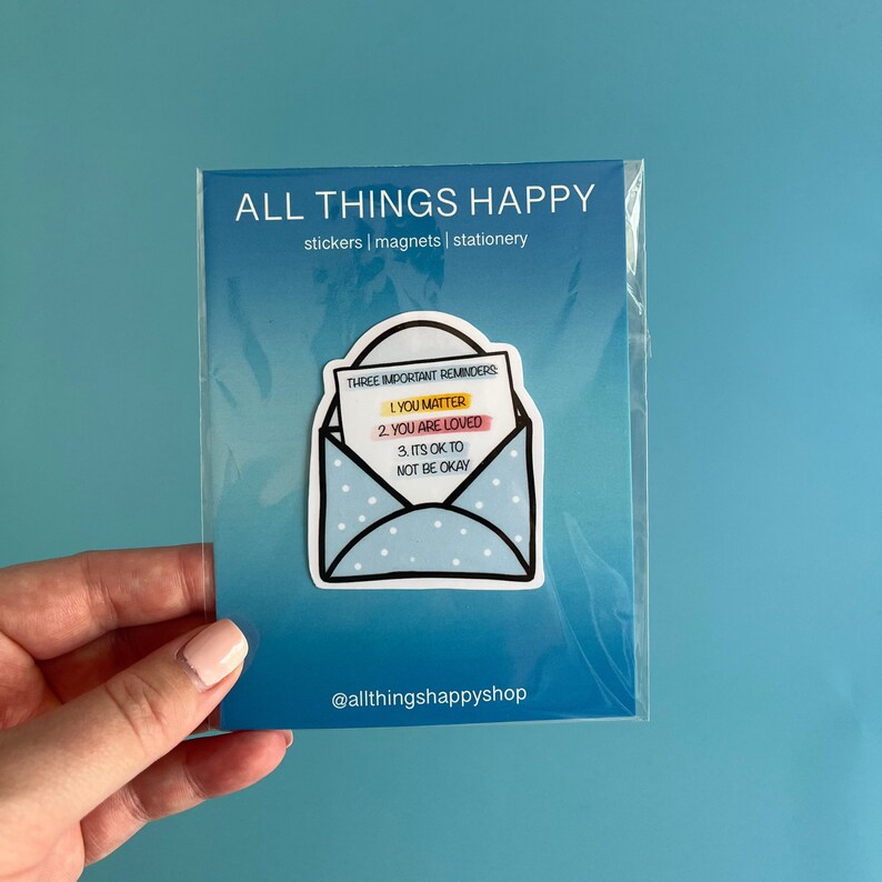 May include: A blue and white envelope sticker with the text "Three Important Reminders: 1. You Matter 2. You Are Loved 3. It's Ok To Not Be Okay" on a white card inside. The sticker is on a white background with the text "@allthingshappyshop" below.