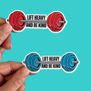 Puede incluir: Dos pegatinas de pesas de barra con el texto "Lift Heavy and Be Kind". Una pegatina tiene pesas rojas y la otra tiene pesas azules.