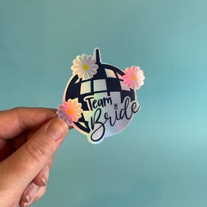 Puede incluir: Una pegatina holográfica con un diseño de bola de discoteca azul marino y la inscripción "Team Bride". La pegatina está adornada con acentos de margaritas rosas y blancas, perfecta para despedidas de soltera o celebraciones nupciales.
