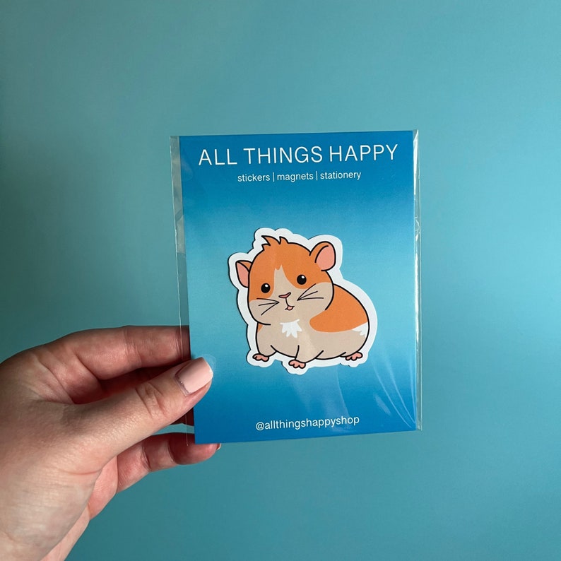 K&ouml;nnte beinhalten: Ein Aufkleber mit einem Cartoon-Hamster mit orangefarbenem und wei&szlig;em Fell. Der Aufkleber befindet sich auf einem blauen Hintergrund mit dem Text "ALL THINGS HAPPY" und "stickers | magnets | stationery" in wei&szlig;. Der Text "@allthingsshappyshop" befindet sich am unteren Rand des Aufklebers.