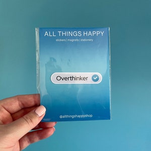 May include: A blue and white sticker with the text "Overthinker" and a checkmark. The sticker is on a blue background with the text "ALL THINGS HAPPY" and "stickers | magnets | stationery" above it. The text "@allthingshappyshop" is at the bottom of the sticker.