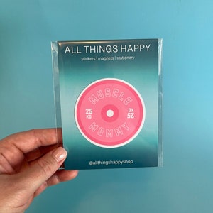 May include: Pink and white vinyl sticker with the text "MUSCLE MOMMY" in a circular weight plate design. The sticker has the text "25 KG" and "SZ 9" on either side of the center circle. The sticker is packaged in a clear plastic sleeve.