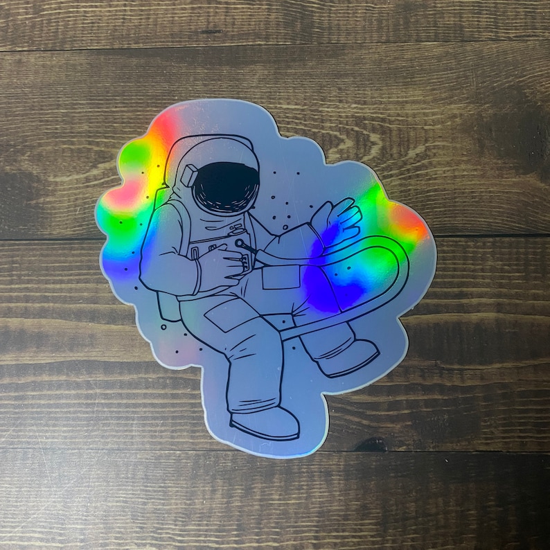 May include: A holographic sticker featuring a black and white line drawing of an astronaut floating in space. The astronaut is wearing a spacesuit and helmet. The sticker has a rainbow iridescent finish.