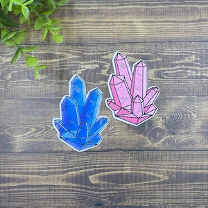 May include: Two crystal stickers, one blue and one pink, with a white background. The crystals are drawn in a cartoon style with a detailed outline.