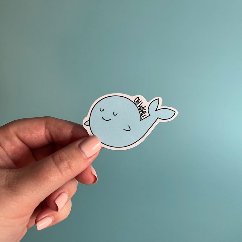 May include: A light blue whale sticker with a smiling face and the text "Oh Whale" on the back fin. The sticker is held in a hand against a light blue background.