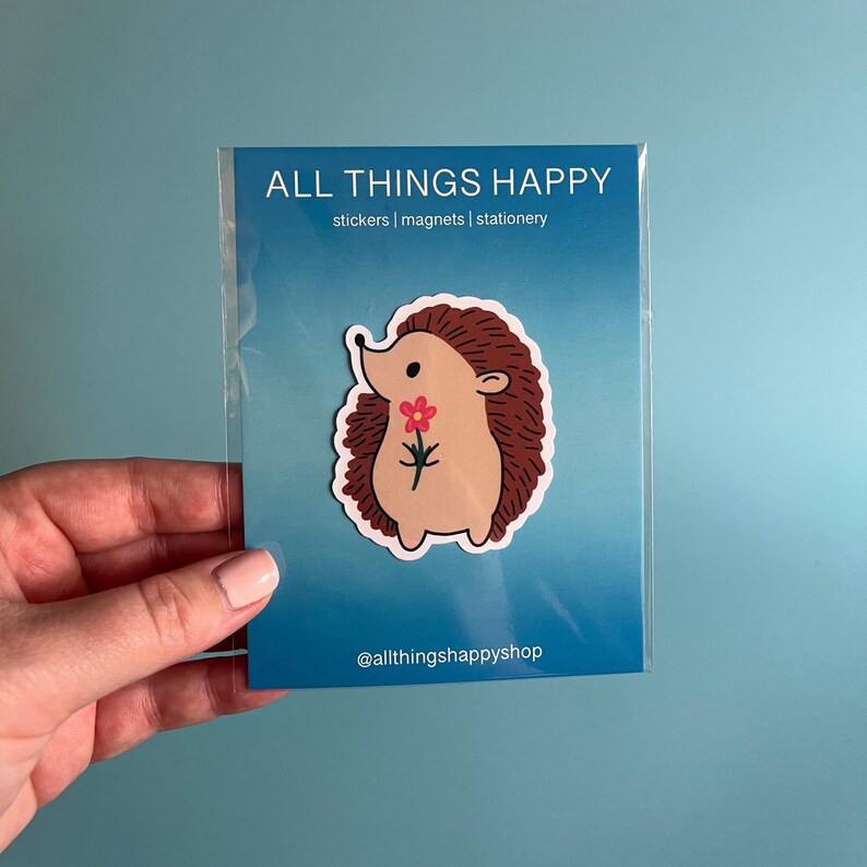 May include: A sticker featuring a cartoon hedgehog holding a pink flower. The sticker is on a blue background with the text "ALL THINGS HAPPY" and "stickers | magnets | stationery" in white. The text "@allthingshappyshop" is at the bottom of the sticker.