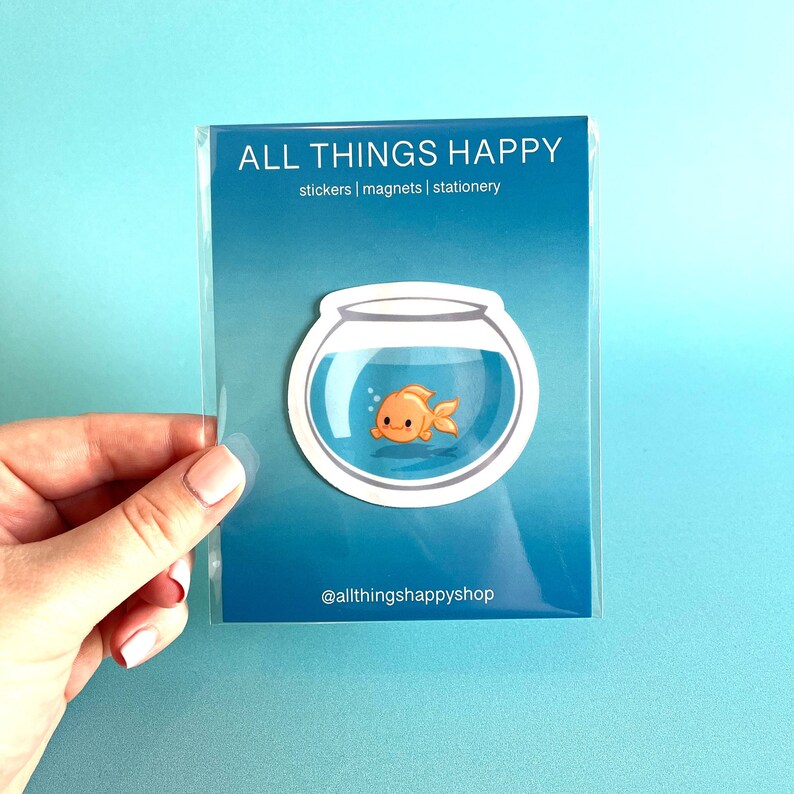 May include: A sticker featuring a cartoon goldfish swimming in a bowl. The sticker is white with a blue background and has the text "ALL THINGS HAPPY" and "@allthingshappyshop" on it.