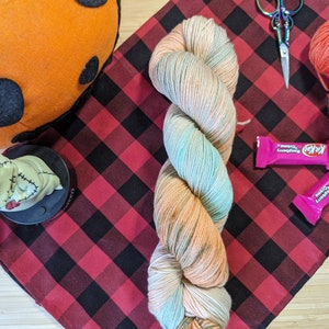 May include: A skein of yarn in shades of peach, teal, and cream, with a black and red checkered fabric background. There are also candy bars, a pumpkin, and scissors in the image.