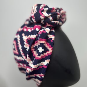 May include: A patterned headband in navy, pink, and white, displayed on a black mannequin head. The fabric features a geometric design. The headband is knotted at the top, creating a stylish accessory.