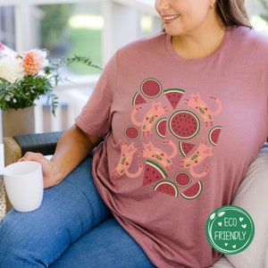 May include: A pink t-shirt with a graphic design of pink cats and watermelon slices. The text "Eco Friendly" is on a green circle in the bottom right corner of the image.