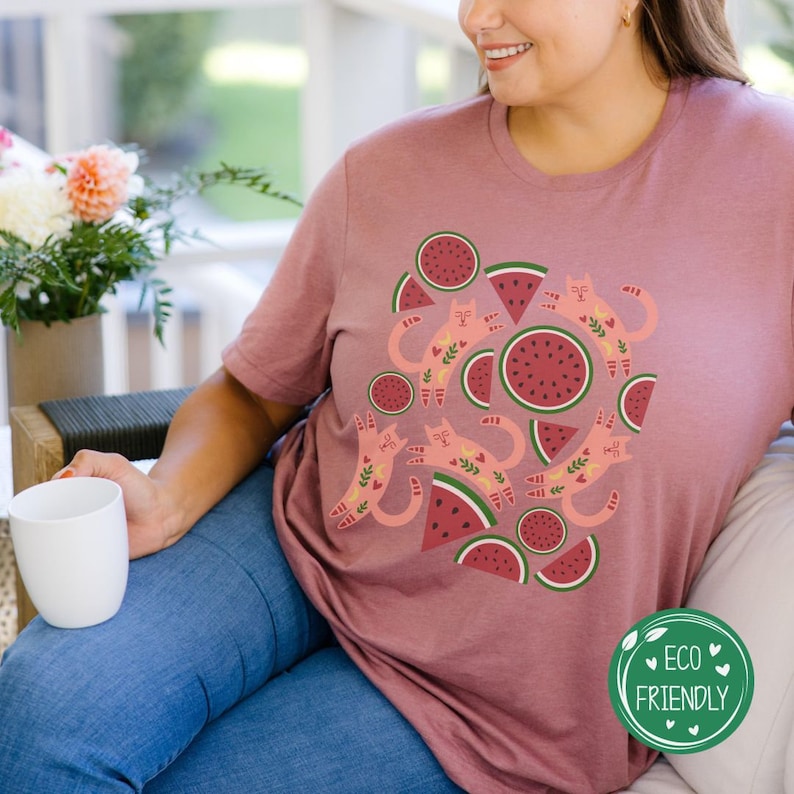 May include: A pink t-shirt with a graphic design of pink cats and watermelon slices. The text "Eco Friendly" is on a green circle in the bottom right corner of the image.