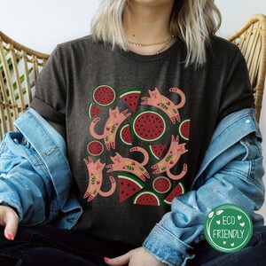 May include: A dark gray t-shirt with a graphic print of pink cats and red watermelon slices. The cats are stylized and have floral details. The watermelon slices are arranged in a circular pattern.