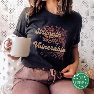 May include: A dark grey t-shirt featuring a brown tree graphic and the text "Strength is being Vulnerable". The shirt is being worn by a person sitting on a white chair. The background is a white wall with a geometric pattern.