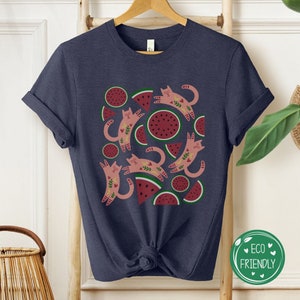 May include: A navy blue t-shirt with a pattern of pink cats and watermelon slices. The cats are stylized and have floral details. The watermelon slices are green and red with black seeds.