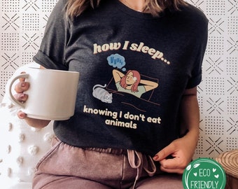 Puede incluir: Una camiseta gris oscuro con un gráfico de una persona durmiendo en la cama con un gato. El texto en la camiseta dice "how I sleep... knowing I don't eat animals".