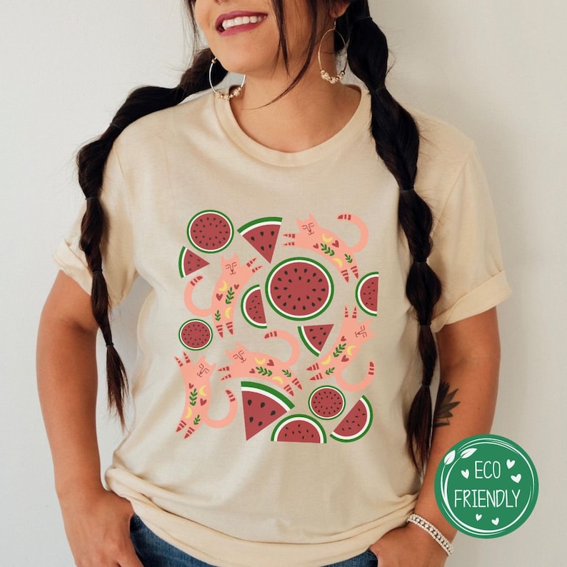May include: A beige t-shirt with a graphic print of pink watermelon slices and stylized cats. The text "Eco Friendly" is printed on a green circle with white hearts.