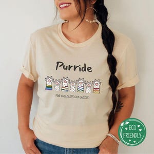 May include: A beige t-shirt with the text "Purrride" and a line of cartoon cats with rainbow paw prints. The text below the cats reads "For Childless Cat Ladies".