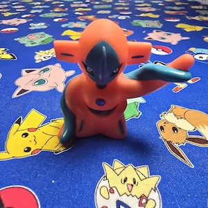 以下が含まれることがあります： オレンジと青色のポケモンキャラクター、デオキシスのフィギュアが、様々なポケモンキャラクターが描かれた青色の生地の上に立っています。フィギュアは特徴的な頭の形と、伸ばされた腕を持っています。背景にはピカチュウ、イーブイ、その他のキャラクターが描かれています。
