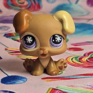 Puede incluir: Una figura de cachorro marrón con ojos grandes y expresivos con diseños de copos de nieve. El perro de juguete tiene un cuerpo marrón claro, orejas marrones oscuras y una oreja marrón claro. El cachorro está sentado sobre una superficie rosa con un patrón colorido.