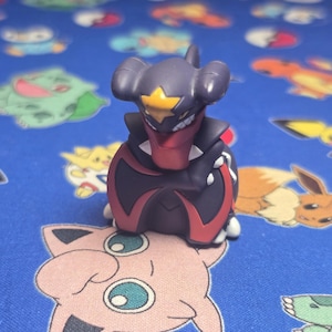 Puede incluir: Una pequeña y detallada figura del personaje Pokémon Garchomp. La figura es principalmente gris oscuro con detalles en rojo y amarillo. Se encuentra sobre una superficie azul decorada con otros personajes Pokémon.