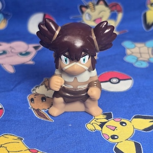 May include: A brown figurine of a character with dark hair styled in two buns. The figure has a white face covering and a brown outfit. The background is a blue fabric with various cartoon character designs.