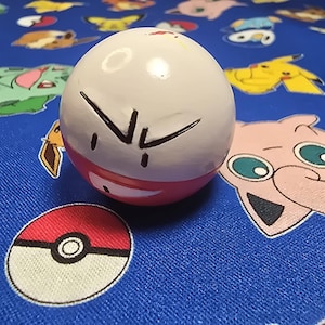 Puede incluir: Una esfera pintada de blanco y rojo con un diseño de cara negra, que se asemeja a un personaje de dibujos animados. Una bola roja y blanca está en primer plano. El fondo es una tela azul con diseños de personajes de dibujos animados.