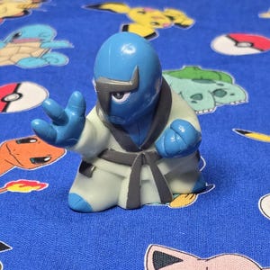 May include: A blue and white action figure of a martial arts character in a fighting pose. The figure has a blue head and hands, a white gi, and a black belt. The background is a blue fabric with cartoon character designs.