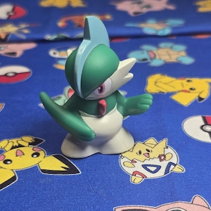 Może przedstawiać: Zielona i biała figurka Gallade, postaci Pokémon, z niebieskim grzebieniem na głowie i czerwonymi akcentami. Figurka jest ustawiona z wyciągniętymi ramionami, na niebieskim tle z tkaniny z różnymi postaciami Pokémon.