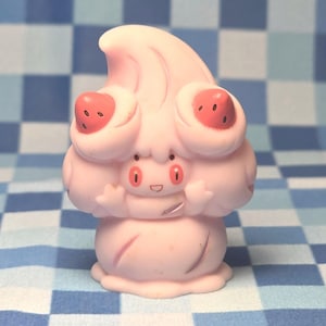Peut inclure: Une petite figurine rose pâle en forme de tourbillon de crème fouettée avec un visage souriant, deux yeux rouges et deux décorations rouges en forme de fraise sur le dessus. Le personnage a de petits bras levés et une base arrondie lisse.
