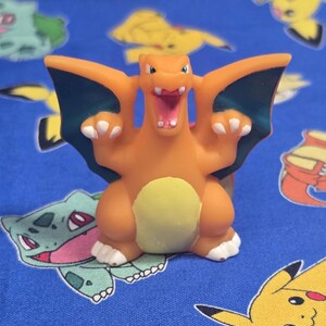 Könnte beinhalten: Orangefarbene Charizard-Figur mit dunkelblauen Flügeln und weißen Krallen, vor blauem Hintergrund mit verschiedenen Pokémon-Charakteren. Die Figur hat ein geöffnetes Maul, das ihre Zähne zeigt.