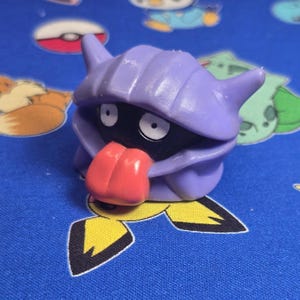 Puede incluir: Una figura de juguete morada con una cara negra, ojos blancos y una lengua roja. La figura tiene un diseño en forma de concha y está sobre una superficie azul estampada con otras figuras de dibujos animados.