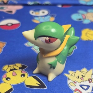 Peut inclure: Figurine Snivy verte et blanche avec des accents jaunes et des yeux rouges. La figurine est sur un fond bleu avec des motifs de personnages de dessins animés. L'objet est un objet de collection.