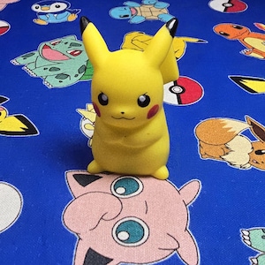 May include: A yellow Pikachu figurine with black-tipped ears, red cheeks, and black eyes. The figurine is on a blue fabric patterned with various Pokémon characters and Poké Balls.