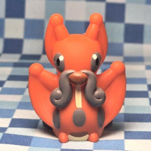 Puede incluir: Una pequeña figura de criatura naranja con grandes orejas puntiagudas, un bigote gris prominente y marcas circulares grises en el cuerpo. La figura tiene ojos negros con reflejos blancos y una pequeña nariz marrón en una pose simétrica.