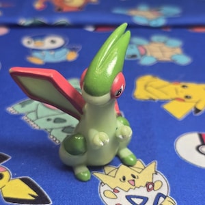 Puede incluir: Una figura verde y roja de un personaje de dibujos animados con grandes alas y una cabeza puntiaguda. La figura tiene un cuerpo verde claro, alas rojas y detalles rojos alrededor de los ojos. El fondo es una tela azul con diseños de personajes de dibujos animados.