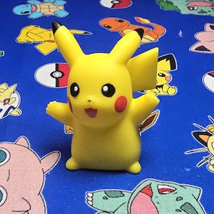 May include: A yellow Pikachu figurine with black-tipped ears, red cheeks, and a smiling expression. The figurine is standing on a blue fabric patterned with various cartoon characters.